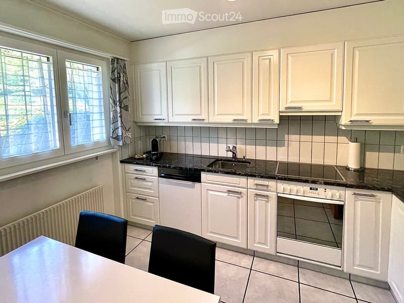 Bright kitchen with white cabinets, black granite countertops, and stainless steel appliances including an oven, dishwasher, and sink. There is a small dining area with a table and two chairs.