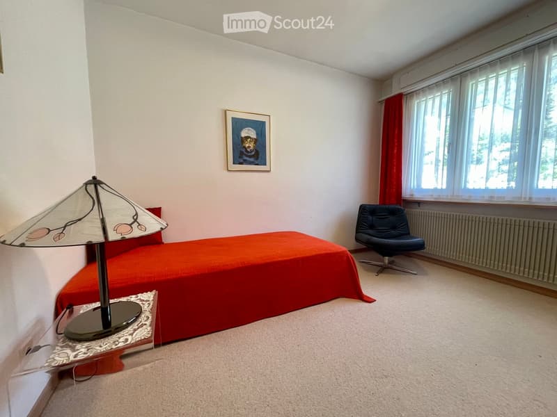 A small bedroom with a red bedspread, a black leather chair, a decorative table lamp, and a framed picture on the wall. The room has large windows with red curtains and a radiator below them.
