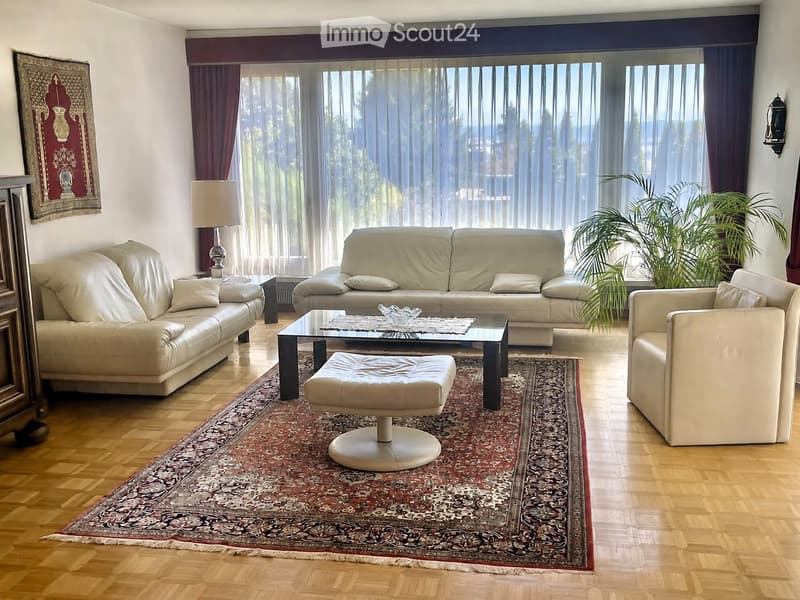 Elegant living room with light-colored leather sofas, a glass coffee table, an ottoman, an armchair, and a large area rug. The room features large windows with sheer curtains and a floor lamp.