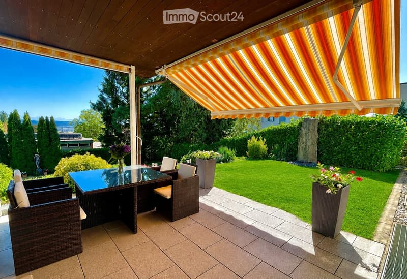 A well-maintained outdoor patio with a glass-top table, wicker chairs, and potted plants, featuring a retractable awning and a view of a green lawn and trees.