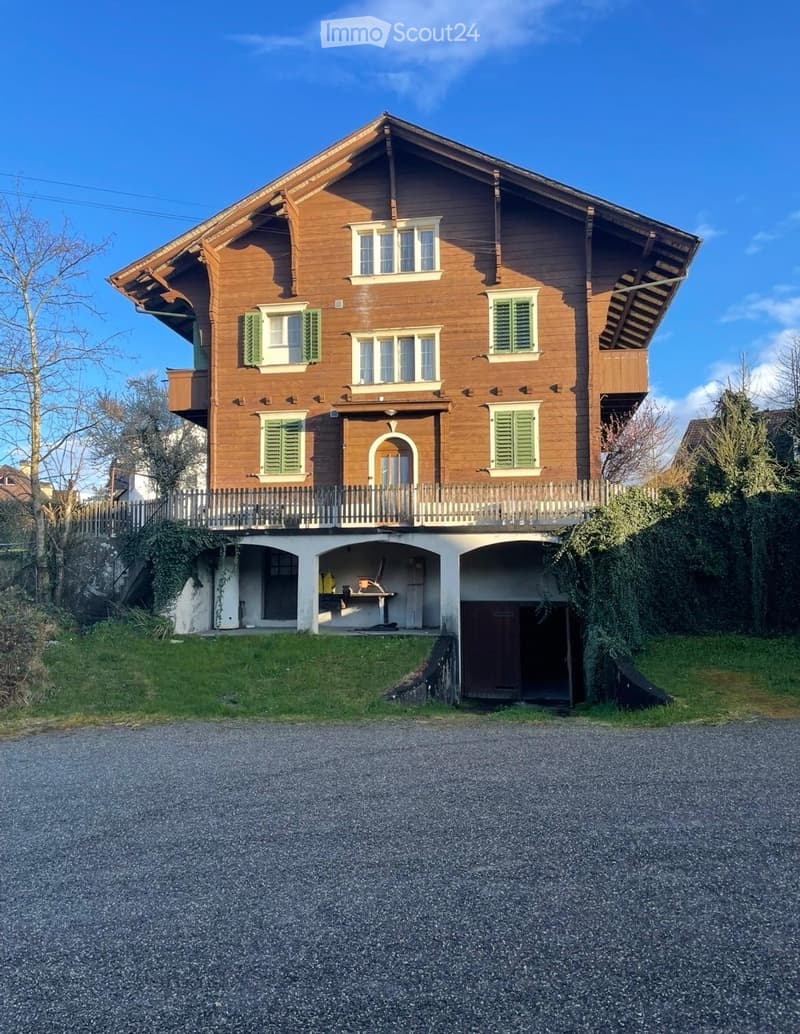 A two-story wooden house with green shutters, a balcony, and a small garage. The house features a pitched roof and is surrounded by a well-maintained garden.