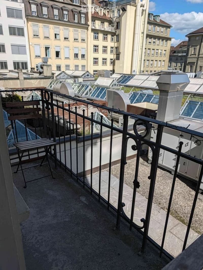 Balcony with metal railing, small wooden chair, overlooking a cityscape with multi-story buildings and a rooftop area.