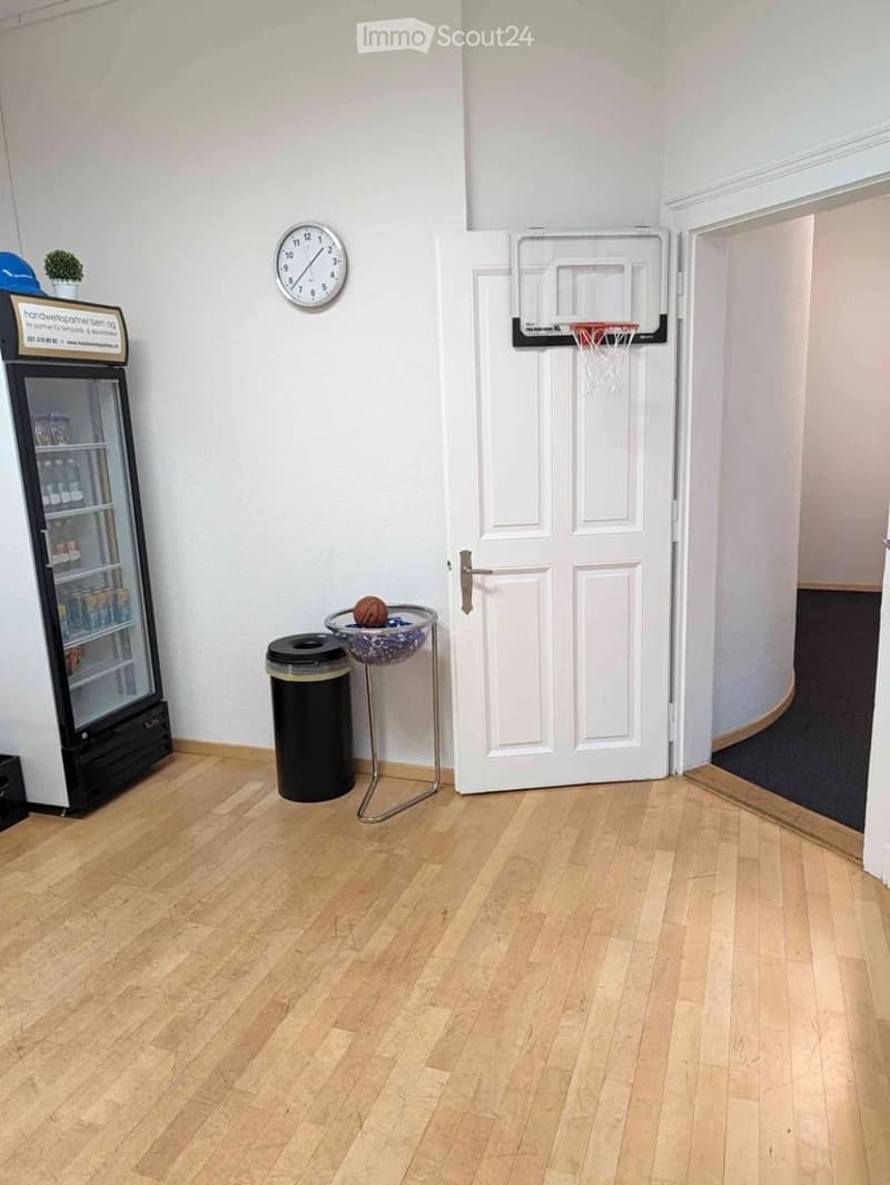 A small, furnished room with light wooden flooring, a white door, a small refrigerator, a clock on the wall, a trash can, a stool with a bowl and a basketball, and a small basketball hoop mounted on the wall.