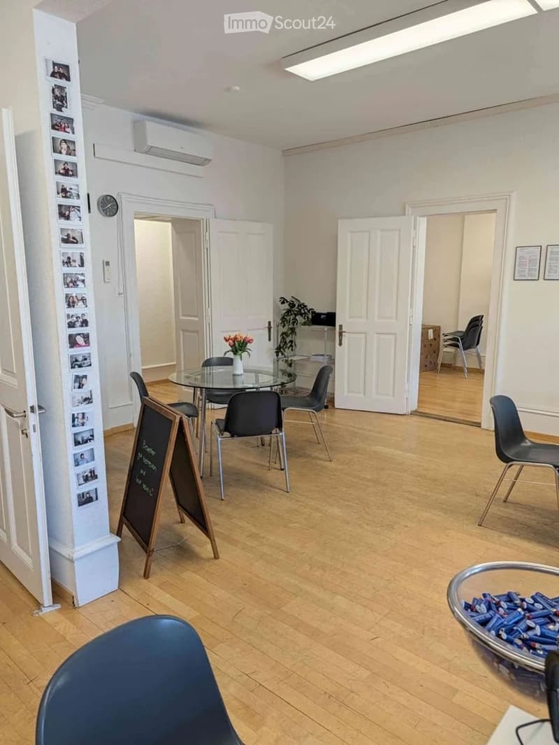 Bright, modern room with light wooden flooring, white walls, and large windows. Features a round glass table with four chairs, a vase with flowers, a clock on the wall, and a screen displaying images.
