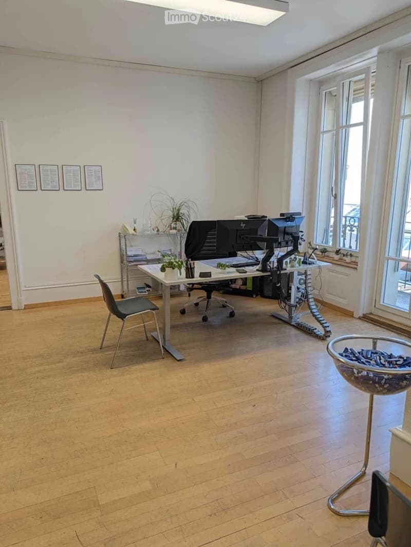 Office space with wooden flooring, large windows, a desk with a computer setup, a chair, and a small plant on the desk.