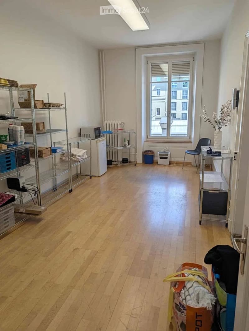 The room features wooden flooring, white walls, and large windows with blinds. It contains metal shelving units with various items, a small refrigerator, a chair, a table with a vase of flowers, and a microwave. There is also a radiator under the window a
