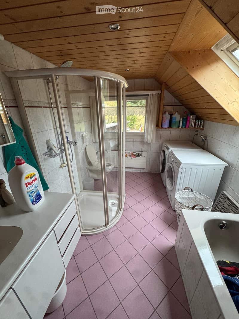 Bathroom with pink tiles, a toilet, a shower, a sink, a washing machine, and a detergent bottle on the counter.