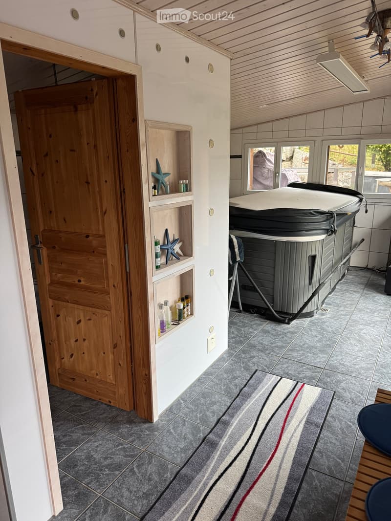 Bathroom with a bathtub, wooden shelves, a mirror, and a small stool. The room has tiled flooring and a striped rug.