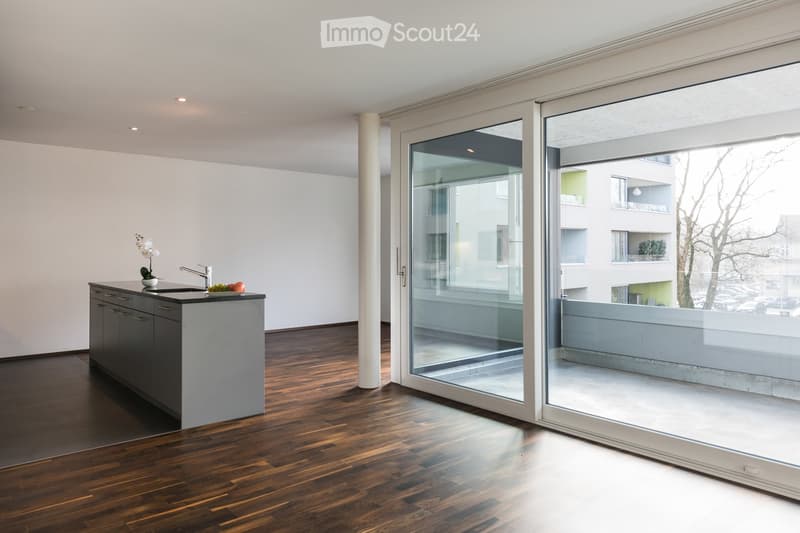 Modern kitchen with dark wooden flooring, sleek grey cabinets, a central island with a sink and faucet, and a vase with white flowers on the counter. Large glass sliding doors open to a balcony area.