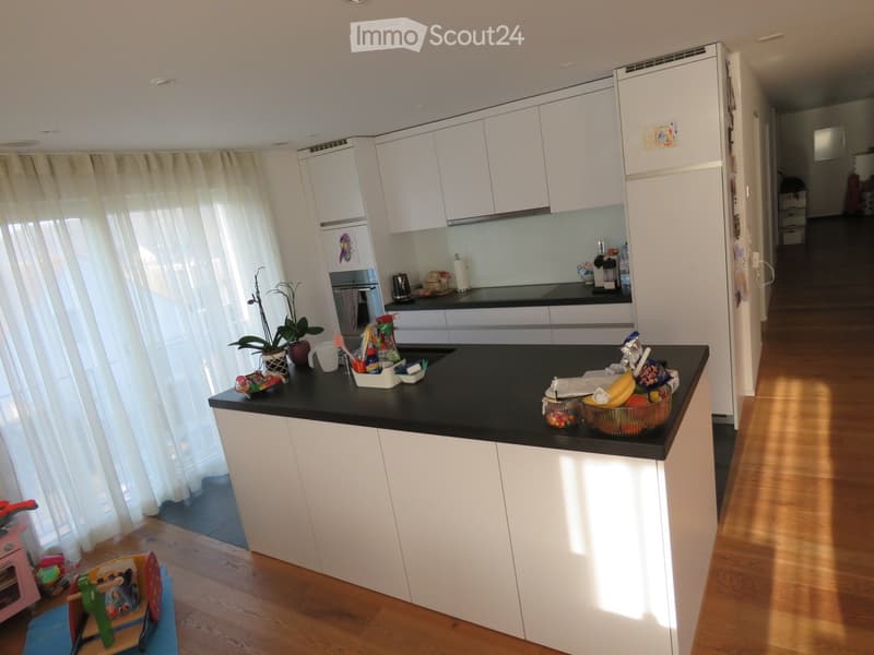 Modern kitchen with white cabinets, black countertops, and a central island. Visible items include a refrigerator, oven, plant, bowl of fruit, and various kitchen accessories.