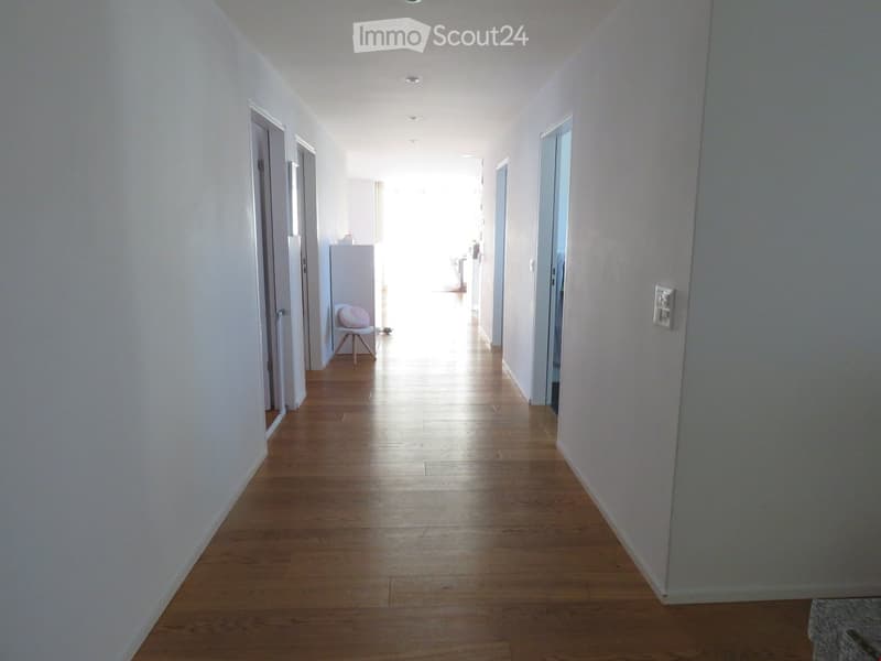 Bright, clean hallway with wooden flooring and white walls, featuring several doors and a small chair with a cushion in the background.