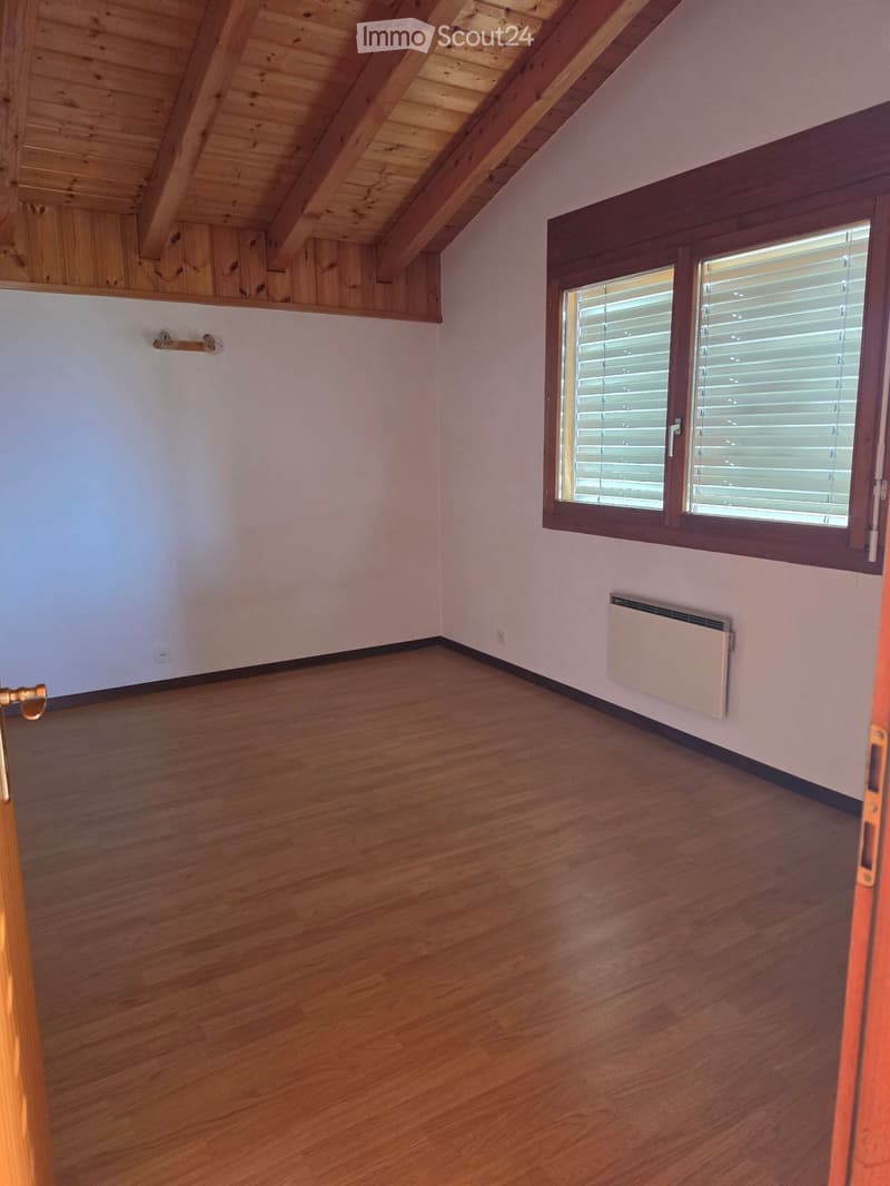 Empty room with wooden flooring, white walls, a window with blinds, and a radiator. The ceiling has exposed wooden beams.