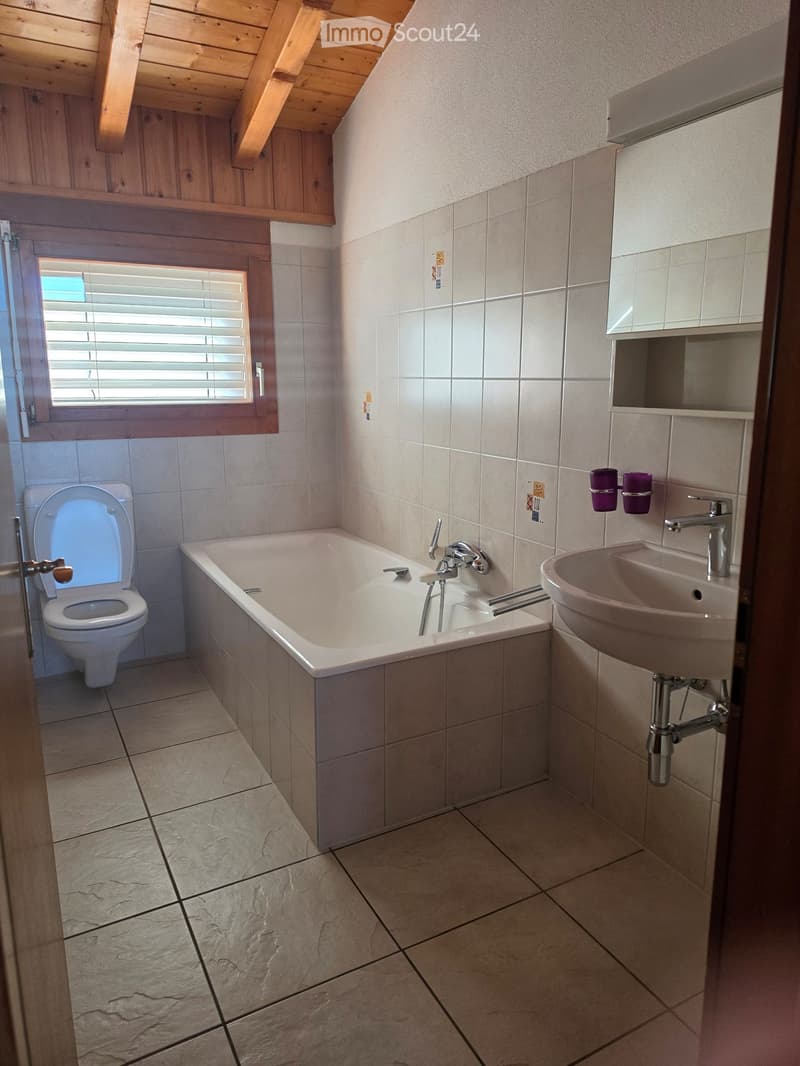 Bathroom with a toilet, bathtub, sink, and a window with blinds. The walls are tiled, and the floor is made of stone tiles.