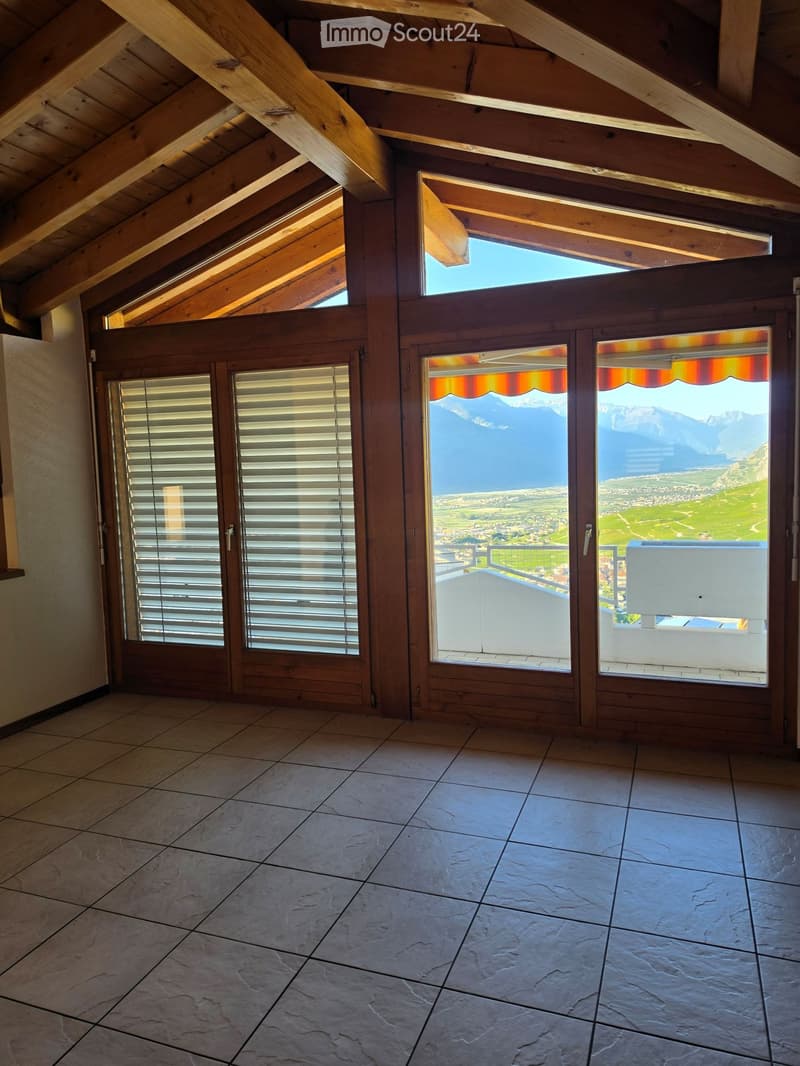 An unfurnished attic room with exposed wooden beams, large windows, and sliding doors leading to a balcony with a scenic mountain view.