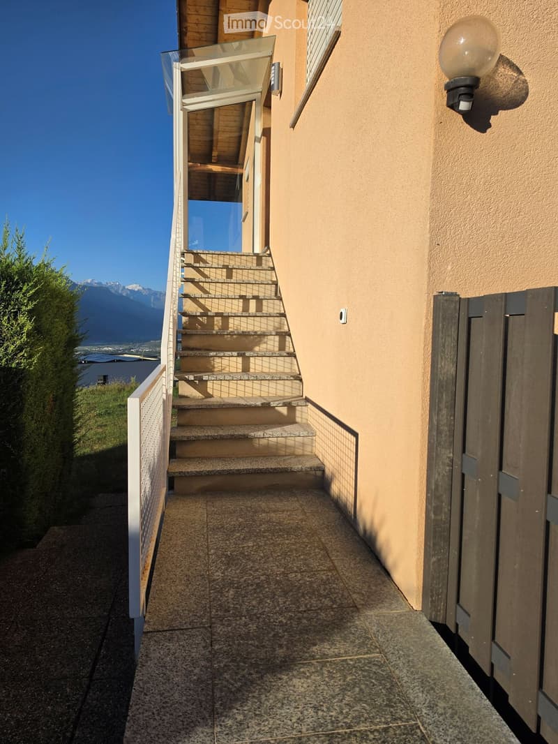 A narrow outdoor corridor with tiled flooring, a staircase leading upwards, and a glass-enclosed balcony area. The walls are light brown, and there is a wall-mounted light fixture on the right side.
