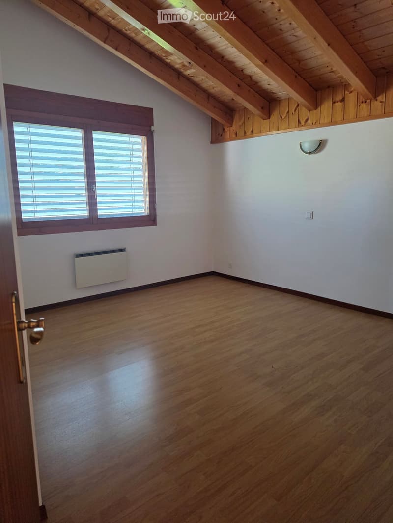Empty attic room with wooden floors, white walls, exposed wooden beams, and a large window with blinds. Features a radiator and a wall-mounted light fixture.