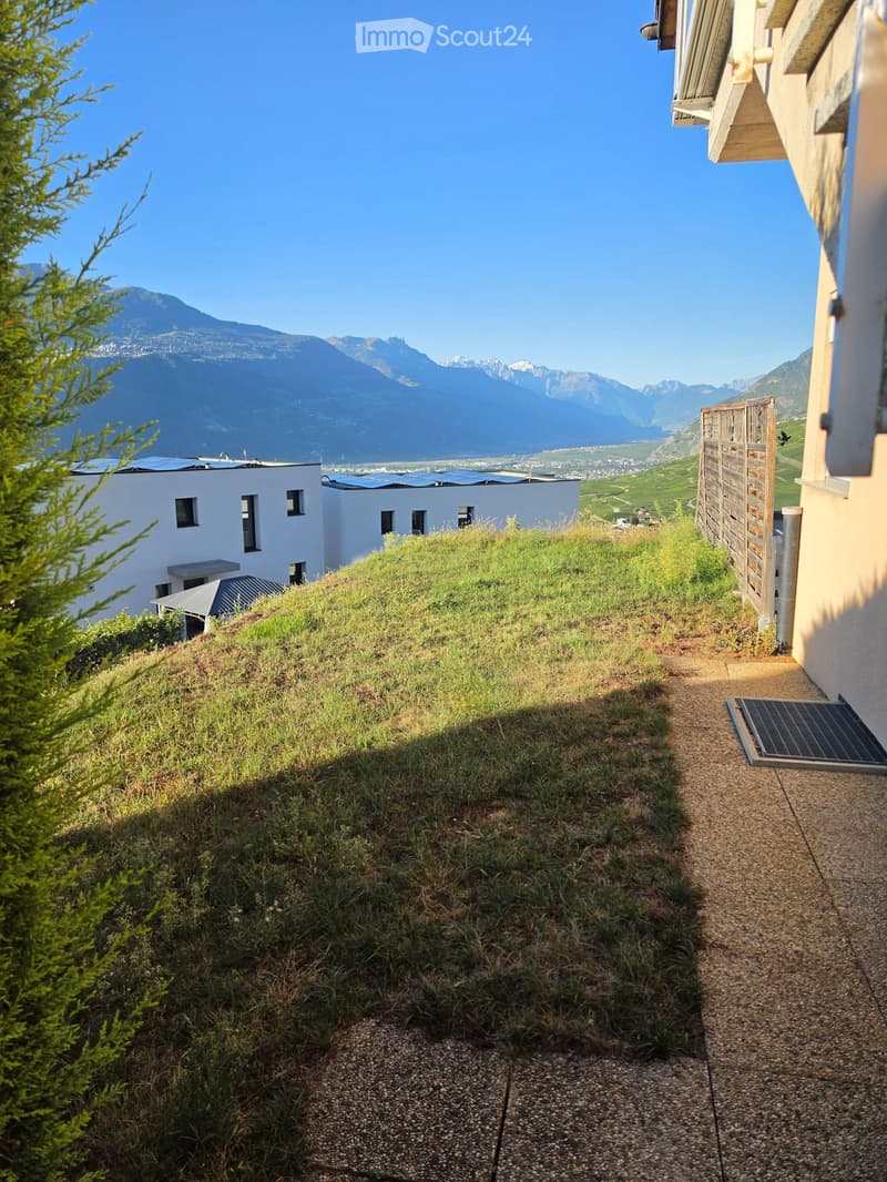 A green roof area with a pathway leading to a building, surrounded by trees and offering a scenic mountain view.