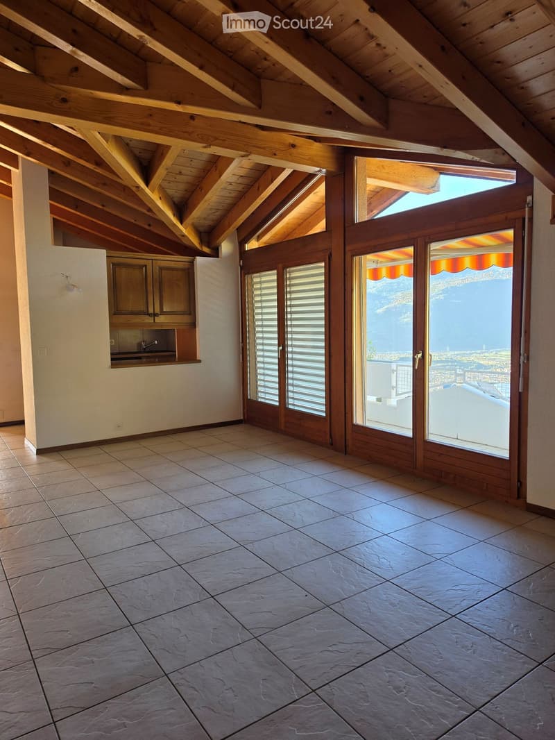 Empty room with exposed wooden beams, tiled floor, and large sliding glass doors leading to a balcony with a scenic view.