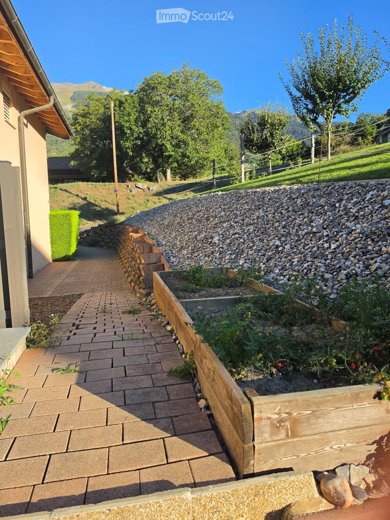 A brick pathway leads to a wooden planter box with plants, surrounded by gravel and a wooden retaining wall, with a green hedge and a view of mountains in the background.