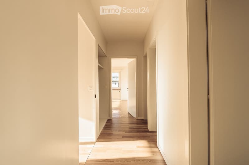 A clean, well-lit hallway with wooden flooring and white walls, featuring several open doors leading to other rooms.