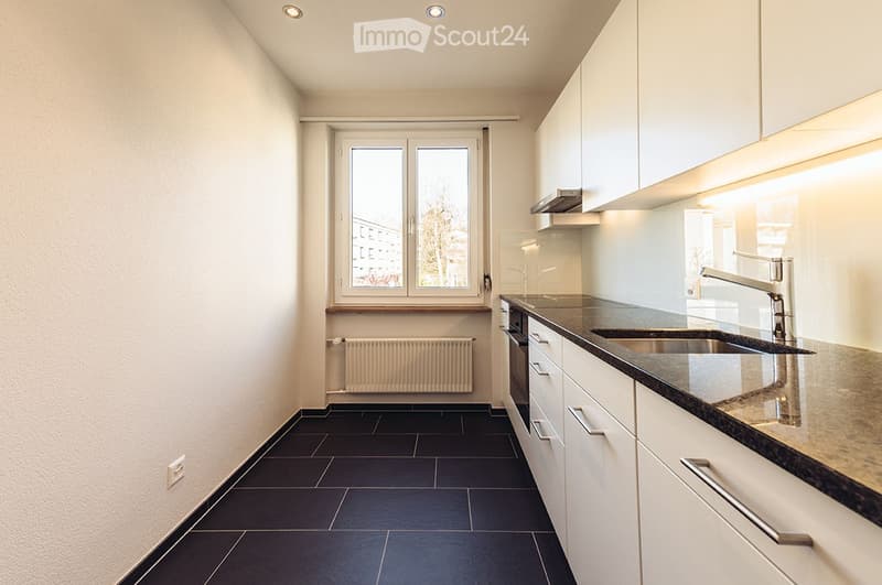 Modern kitchen with white cabinets, dark tiled floor, stainless steel sink, and built-in oven. Features a large window with a view of the outside.