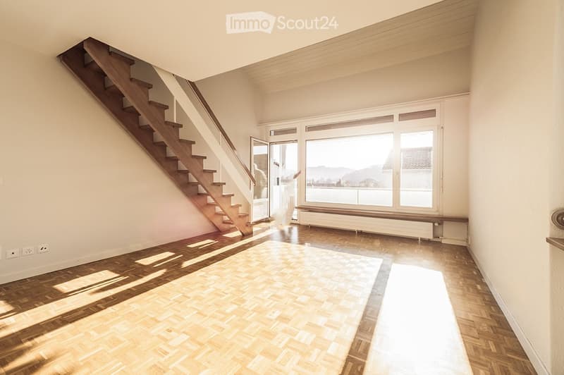 Empty room with wooden flooring, staircase leading upwards, large windows with a view, and a radiator under the windows.