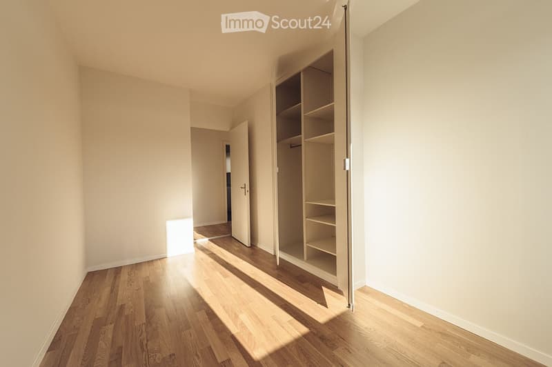 Empty room with wooden flooring, light-colored walls, and an open closet with empty shelves. Sunlight streams in from a window, casting shadows on the floor.
