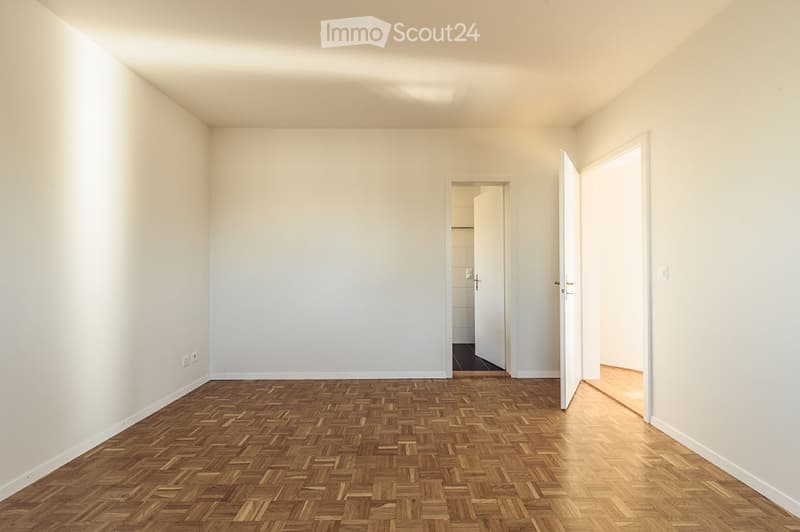 Empty room with wooden herringbone flooring and white walls, featuring two open doors leading to adjacent spaces.