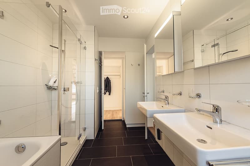 Modern bathroom with a bathtub, two sinks, a large mirror, and a glass-enclosed shower. The space features white tiles, dark flooring, and sleek chrome fixtures.