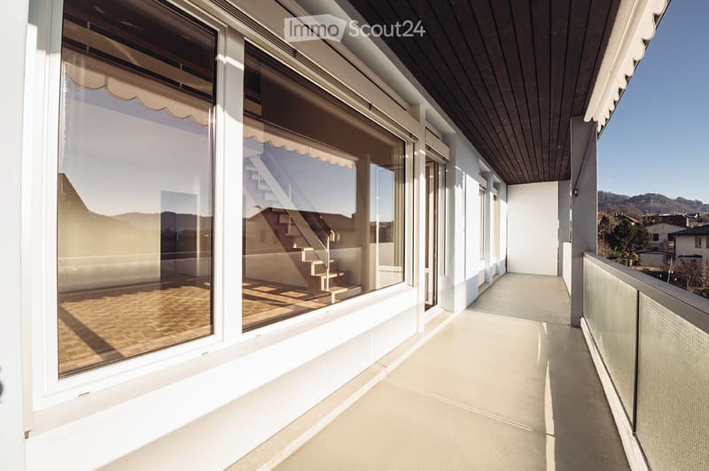 Spacious balcony with large windows, wooden flooring, and a view of the surrounding landscape.
