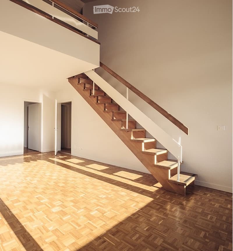 Bright, open hallway with wooden flooring and a staircase leading to an upper level. The space features white walls, a glass railing on the staircase, and natural light streaming in from above.