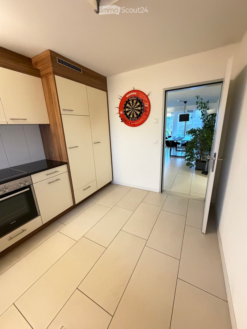 Modern kitchen with white cabinets, black countertop, and a dartboard on the wall. An open doorway leads to a dining area with a table and chairs.