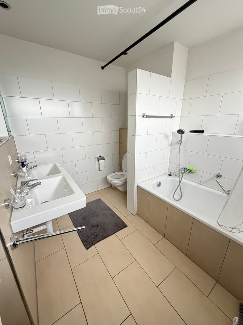 Modern bathroom with white tiles, double sink, toilet, bathtub, and a small dark grey rug on the floor.