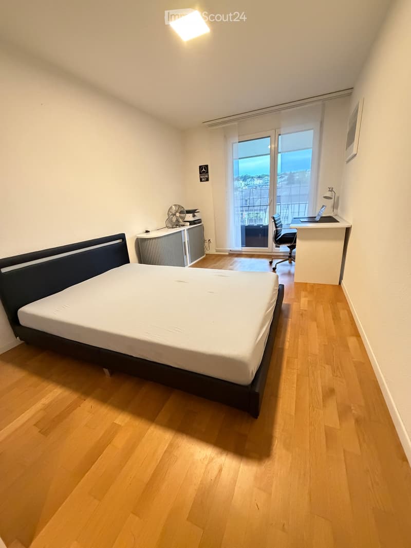 Modern bedroom with wooden flooring, a double bed, a desk with a laptop, a chair, a fan, and a printer. Large windows offer a scenic view.