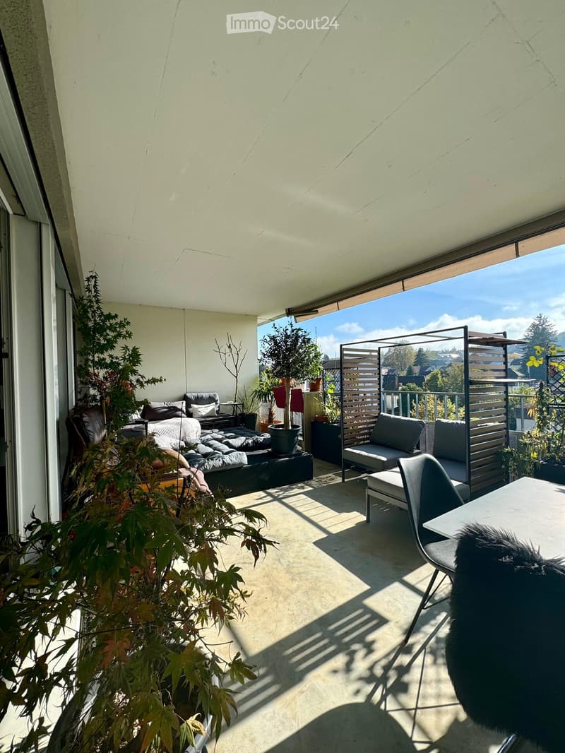 Modern balcony with a view, featuring a sofa, chairs, a table, and potted plants.