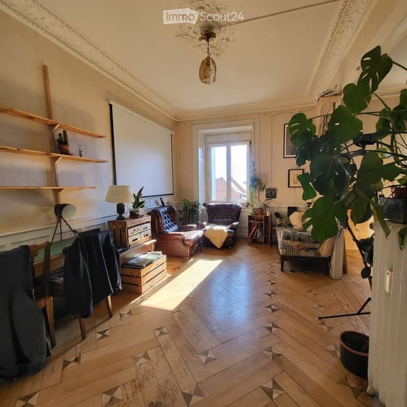 Cozy living room with wooden flooring, large window, sofa, armchair, small table, lamp, shelves, and plants.