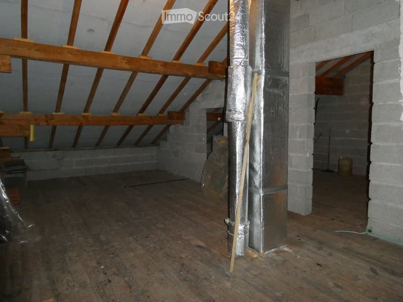 Empty attic space with wooden beams, exposed wooden floor, and insulation on a central pillar. No furniture or equipment present.
