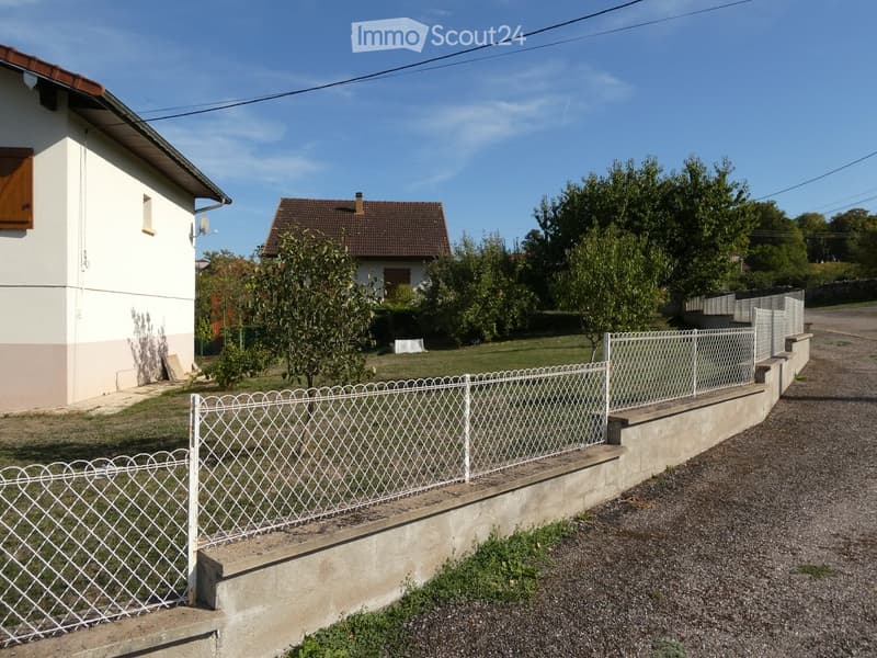 Two-story house with white and beige exterior, red-tiled roof, and a fenced garden area with trees and shrubs.