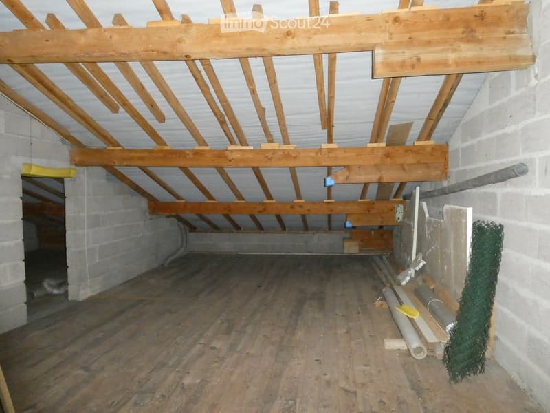 Empty attic space with exposed wooden beams and a concrete floor, showing construction materials and tools.