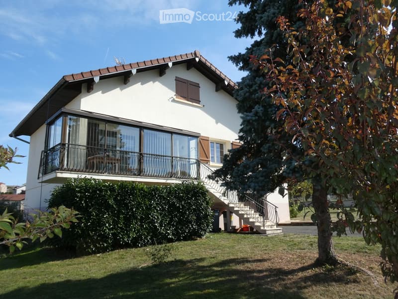 Two-story white house with a brown roof, large windows, and a balcony on the upper floor. The house features wooden shutters and is surrounded by a well-maintained lawn and trees.
