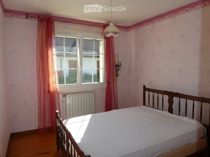 A cozy bedroom with pink floral wallpaper, a wooden bed with a white bedspread, pink curtains, a radiator, and a hanging light fixture.
