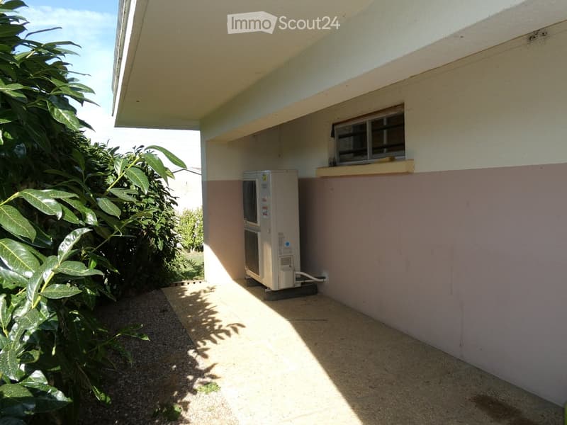 Outdoor area with a covered section, air conditioning unit, window, and surrounding greenery.