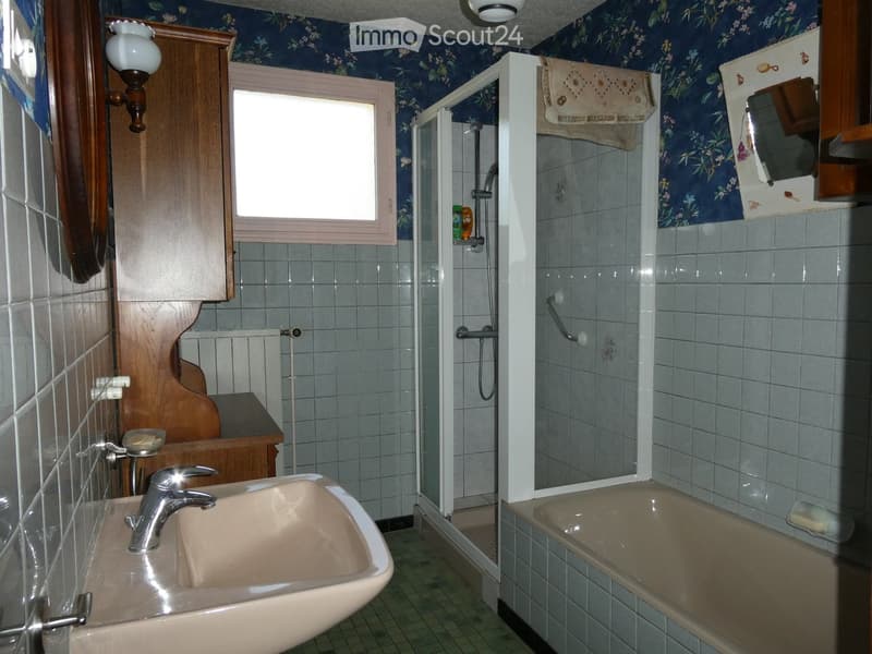 Bathroom with a sink, bathtub, shower, mirror, and wooden cabinet. The walls are tiled with blue floral wallpaper, and there is a window above the sink.