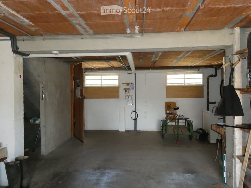 The image shows a partially furnished basement or utility room with exposed wooden beams on the ceiling, concrete floors, and white walls. Visible objects include a small wooden chair, a table, shelves, a door leading to another room, and a small stool. T