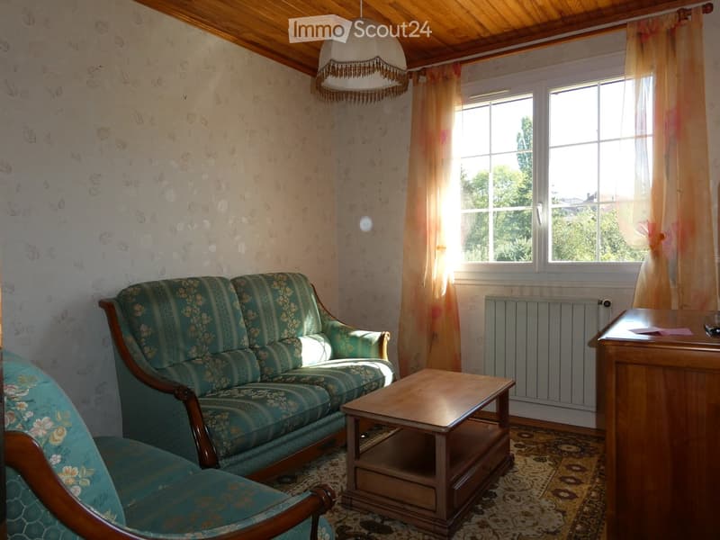 Living room with a green patterned sofa, armchair, coffee table, radiator, and a small wooden table with a lamp on it. The room has wooden beams on the ceiling, patterned wallpaper, and large windows with sheer curtains.