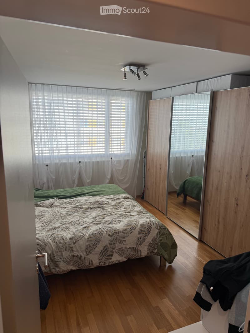 A modern bedroom with a large bed, patterned duvet, wooden wardrobe with mirrored doors, and sheer curtains covering the window. The room features wooden flooring and recessed ceiling lighting.