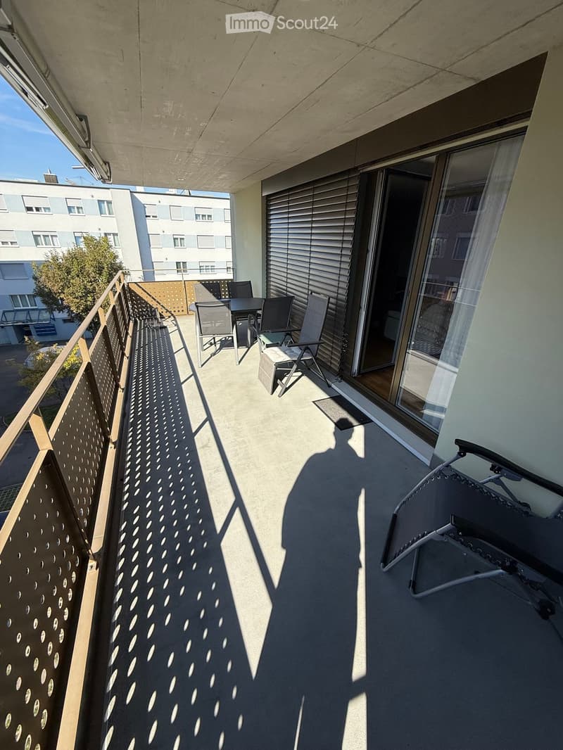Balcony with a table, chairs, and a lounge chair, overlooking a residential area with buildings and trees.