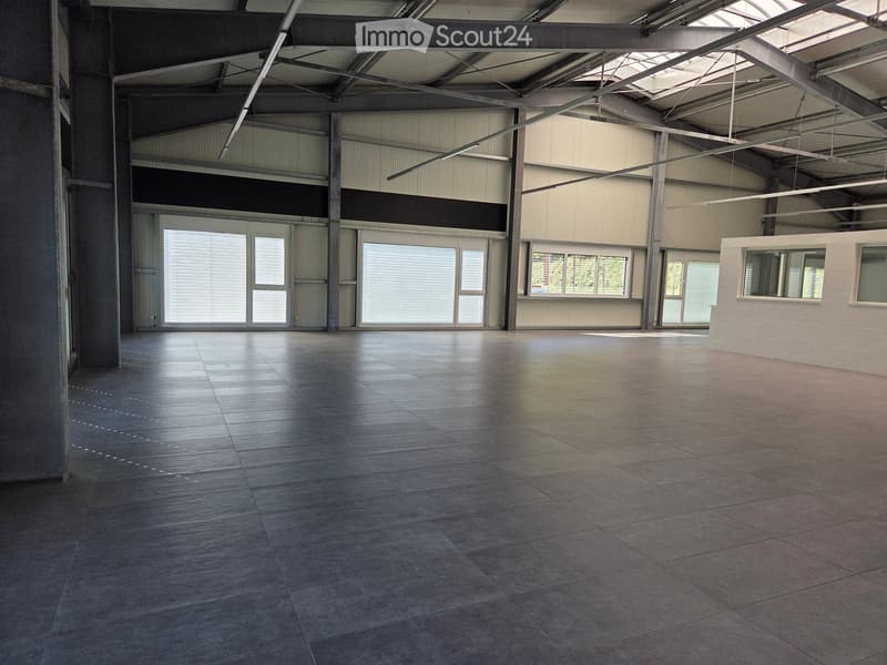 Large, empty industrial-style room with polished concrete floors, exposed metal beams, large windows, and a reception area with a white brick wall and small window.