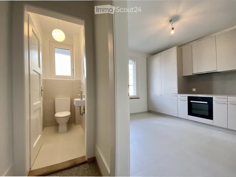 The image shows a modern, minimalist bathroom and kitchen area. The bathroom features a toilet, sink, and a window, while the kitchen has white cabinets, a built-in oven, and a sleek design.
