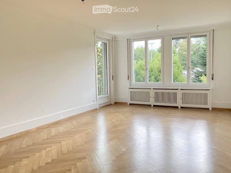 Empty room with light wooden herringbone flooring, white walls, large windows with white frames, and a white radiator unit below the windows.